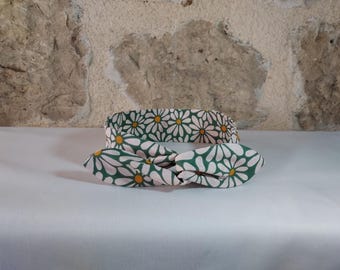 May include: Two fabric headbands with a daisy pattern. The headbands are white daisies with yellow centers on a pink background, set against a dark green background. One headband is a simple band, and the other is a knotted bow.