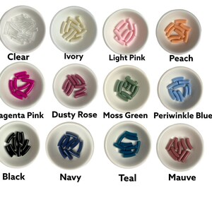 Op de afbeelding: Assortiment van 12 kleine, gebogen kunststof onderdelen in verschillende kleuren, waaronder helder, ivoor, lichtroze, perzik, magenta roze, stoffig roze, mosgroen, periwinkleblauw, zwart, marineblauw, teal en mauve. De onderdelen zijn gerangschikt in kleine witte kommen.