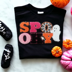May include: A black crewneck sweatshirt featuring a textured chenille patch that spells out the word SPOOKY in orange, pink, and black letters. The letter O is replaced by a spiderweb design, and a white ghost figure is positioned next to the Y.