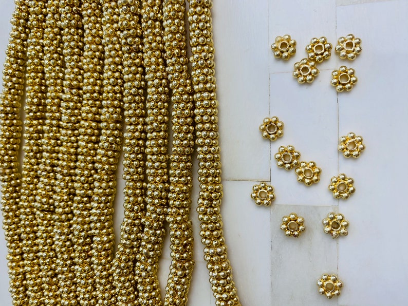 May include: A close-up of a strand of gold beads, with a scattering of gold flower-shaped beads on a white surface.