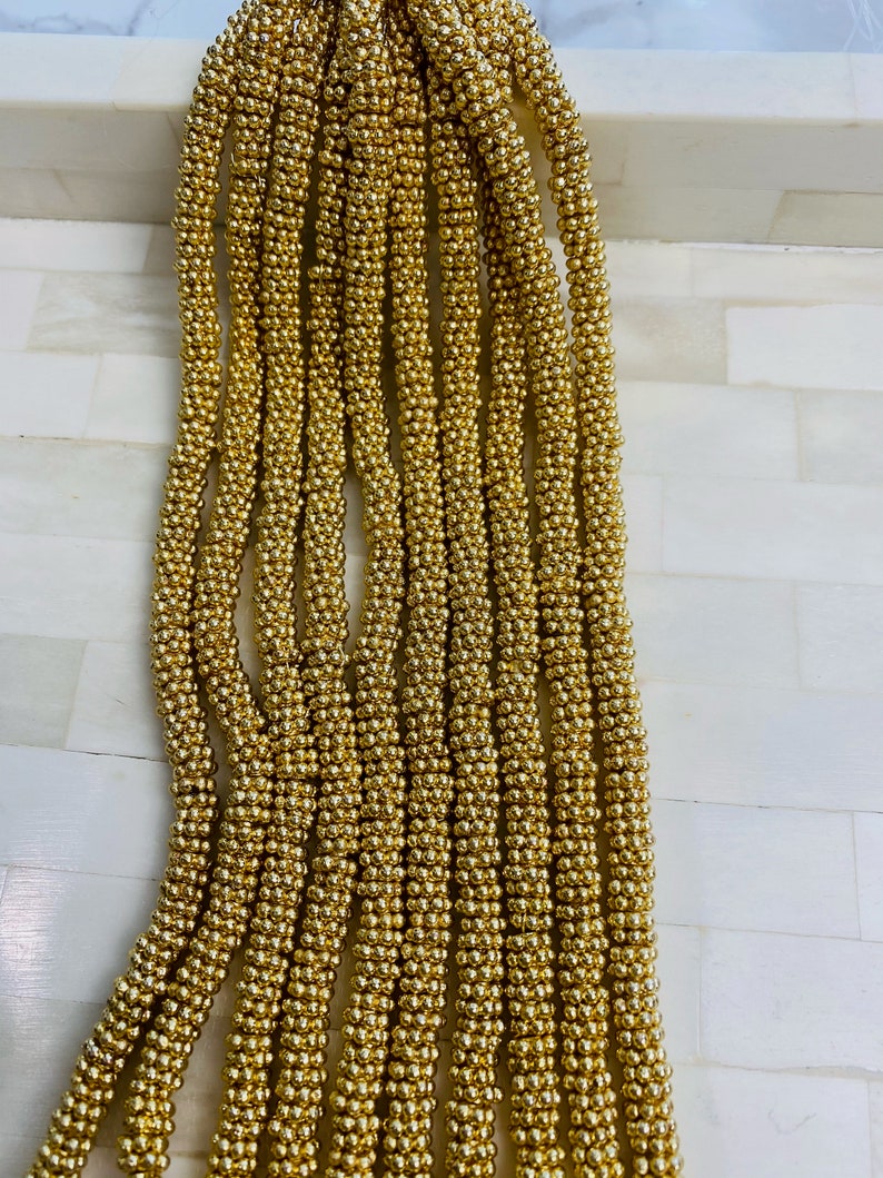 May include: A strand of gold-colored beads, each bead is small and round. The beads are strung together to form a long, continuous strand.