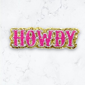 May include: Pink and gold glitter iron-on patch with the word "Howdy" in a bold, cursive font.
