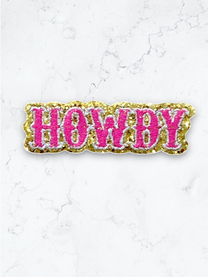 May include: Pink and gold glitter iron-on patch with the word "Howdy" in a bold, cursive font.