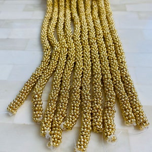 May include: A bundle of gold-colored beaded cords, each cord is made of small, round beads. The cords are arranged in a fan shape, with the ends of the cords pointing towards the bottom of the image.