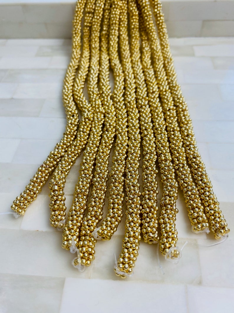 May include: A bundle of gold-colored beaded cords, each cord is made of small, round beads. The cords are arranged in a fan shape, with the ends of the cords pointing towards the bottom of the image.
