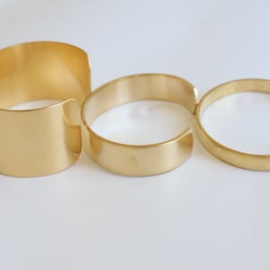 May include: Three gold-colored metal cuff bracelets of varying widths are displayed against a white background. The bracelets are open-ended, with a smooth, polished finish. The largest cuff is wide, while the others are narrower.