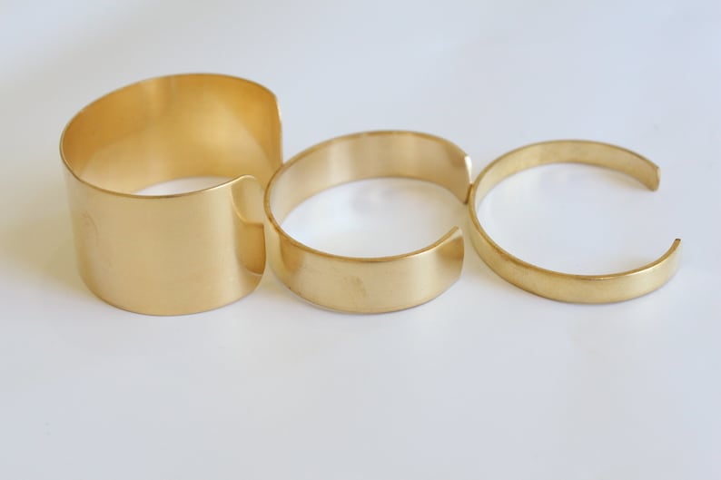 May include: Three gold-colored metal cuff bracelets of varying widths are displayed against a white background. The bracelets are open-ended, with a smooth, polished finish. The largest cuff is wide, while the others are narrower.