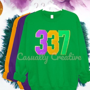 Peut inclure: Sweat-shirt vert avec le numéro 337 en paillettes violettes, dorées et vertes. Le texte "Casually Creative" est imprimé sous les chiffres.