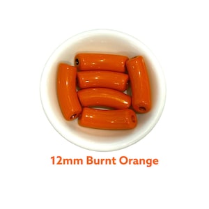 May include: A white bowl contains several burnt orange barrel beads, each about 1.2 cm long. The beads are glossy and cylindrical with rounded ends. The text "12mm Burnt Orange" is displayed below the bowl.