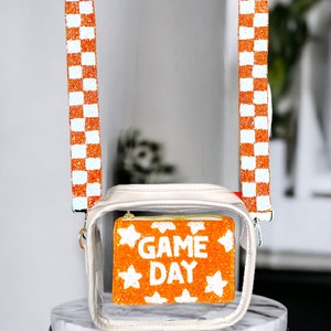 May include: Clear plastic crossbody bag with a white and orange checkered strap. The bag has a clear front pocket with a beaded orange and white "GAME DAY" design.