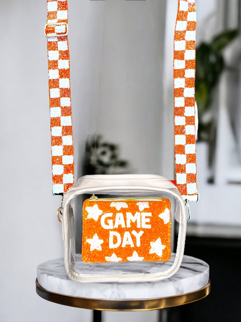 May include: Clear plastic crossbody bag with a white and orange checkered strap. The bag has a clear front pocket with a beaded orange and white "GAME DAY" design.