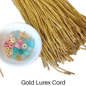 May include: A bundle of gold lurex cords with metallic gold aglets positioned next to a small white bowl containing a collection of assorted small beads in shades of pink, blue, and white, along with a tiny gold cross and flower charm.