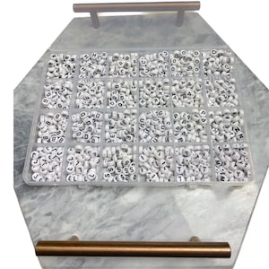 May include: A white plastic storage box with 24 compartments filled with white letter beads. Each compartment contains a different letter of the alphabet.