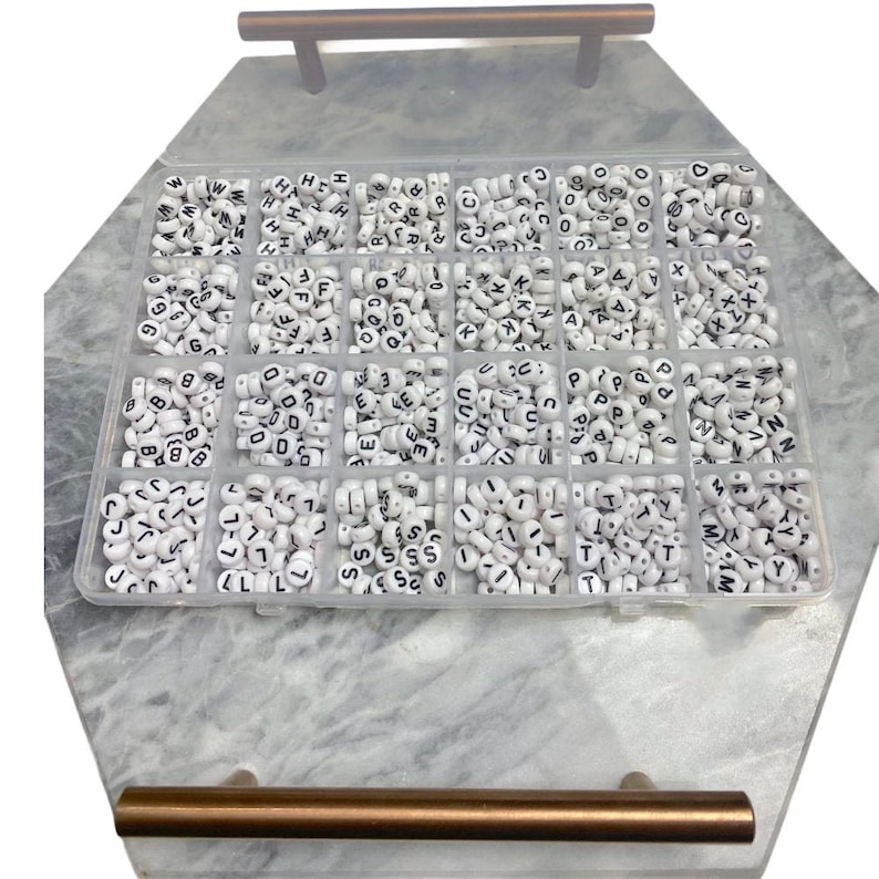 May include: A white plastic storage box with 24 compartments filled with white letter beads. Each compartment contains a different letter of the alphabet.