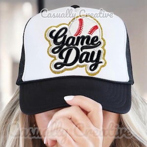 以下が含まれることがあります： 黒のメッシュバックが付いた白いトラッカーハット。ハットの前方には、野球ボールと「Game Day」の文字が刺繍された、金色と黒色のパッチが付いています。