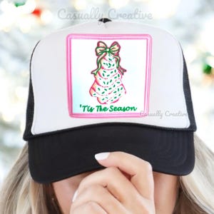 May include: A white and black trucker hat with a pink and green embroidered Christmas tree design. The text 'Tis The Season is embroidered on the hat.