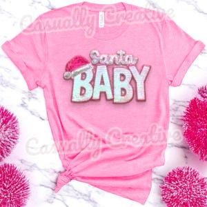 Puede incluir: Camiseta rosa con un diseño brillante blanco y azul "Santa Baby" con un gorro de Papá Noel rojo.