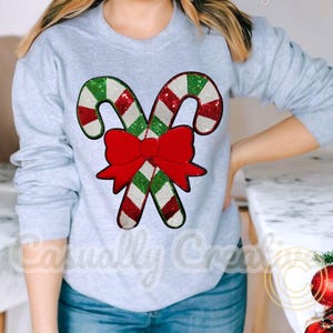May include: A light gray sweatshirt featuring a festive design. The design includes two candy canes with red, white, and green sequins, and a large red bow. The text "Casually Creative" is printed on the sweatshirt.