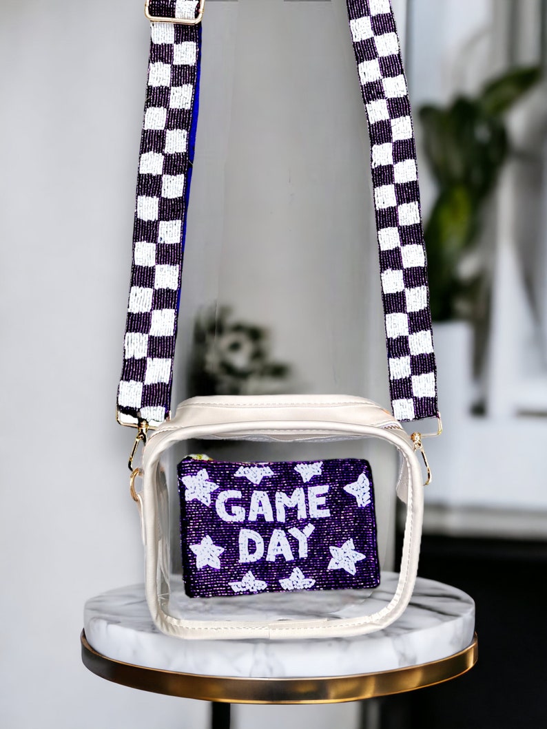 May include: Clear plastic crossbody bag with a white and black checkered beaded strap. The bag has a purple beaded pouch inside with white stars and the text "GAME DAY".