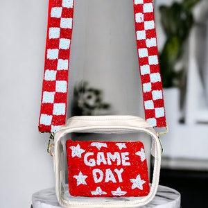 May include: Clear plastic crossbody bag with a red and white checkered beaded strap. The bag has a clear front panel that shows a red beaded clutch with white stars and the text "GAME DAY".