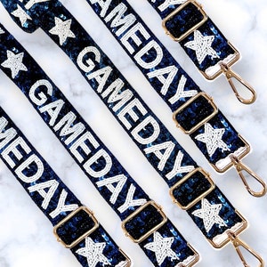以下が含まれることがあります： 金色の金具と白いビーズで「GAMEDAY」と書かれた、青と白のキラキラしたストラップが4本。ストラップは白い星で飾られています。