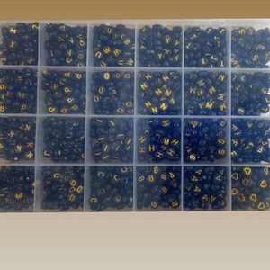 May include: A clear plastic storage box filled with small black beads with gold letters on them. The beads are organized in rows and columns. The letters of the alphabet are visible.