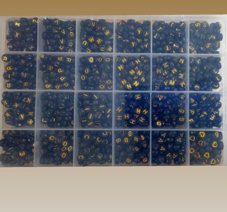 May include: A clear plastic storage box filled with small black beads with gold letters on them. The beads are organized in rows and columns. The letters of the alphabet are visible.