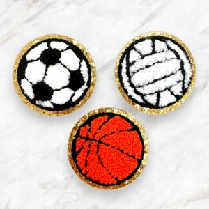 Puede incluir: Tres posavasos redondos, con borde dorado, cubiertos de fieltro con diferentes diseños de pelotas de deporte. Los posavasos presentan un balón de fútbol, un balón de voleibol y un balón de baloncesto.