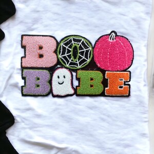 May include: White t-shirt with a colorful embroidered design that says "BOO BABE". The design includes a ghost, a spiderweb, and a pumpkin.