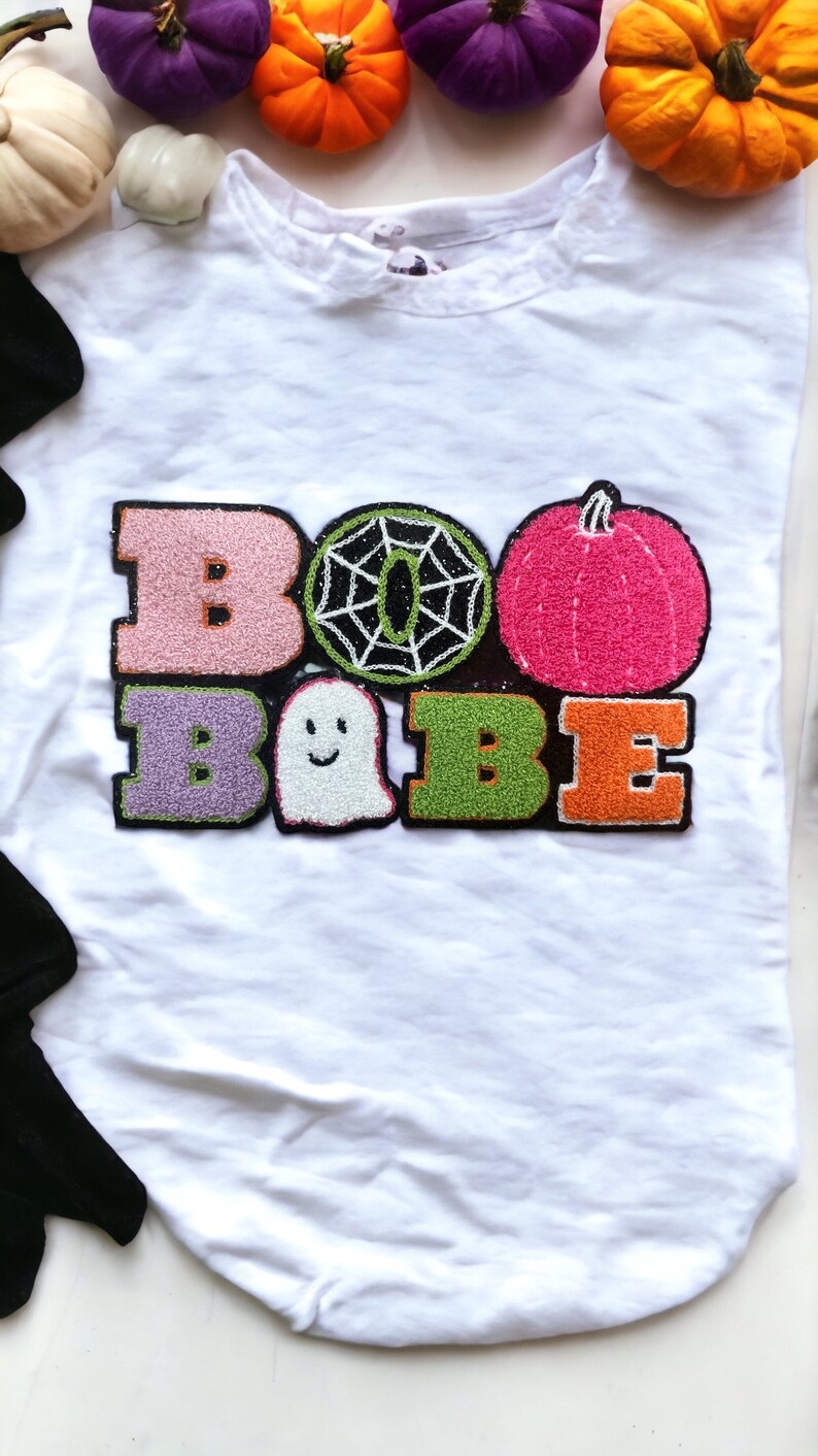 May include: White t-shirt with a colorful embroidered design that says "BOO BABE". The design includes a ghost, a spiderweb, and a pumpkin.