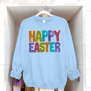 Könnte beinhalten: Hellblauer Pullover mit den Worten "Frohe Ostern" in Regenbogenfarben mit Glitzerakzenten.
