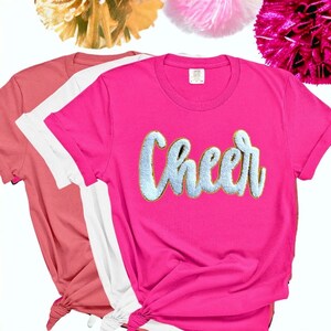 May include: Three t-shirts in pink, white and coral with the word "Cheer" in gold glitter lettering on the pink shirt.