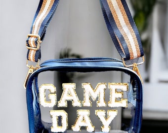 May include: Clear blue and white crossbody bag with a gold zipper and strap. The bag has a clear front with the words "GAME DAY" in gold and white letters.