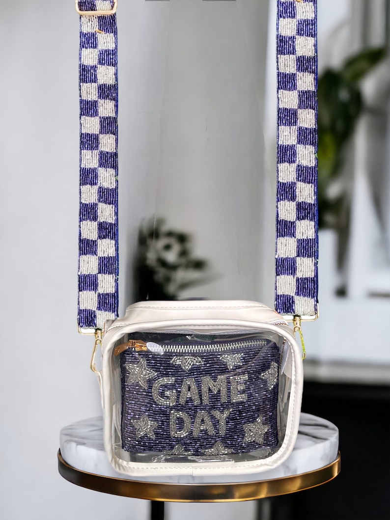 May include: Clear crossbody bag with a white base and a clear front panel. The front panel features a beaded design with the words "GAME DAY" in silver beads. The bag has a blue and white checkered beaded strap.