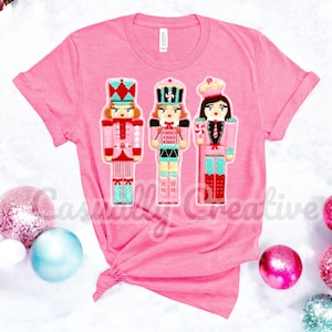 Könnte beinhalten: Pinkes T-Shirt mit einem Motiv von drei Nussknackerfiguren. Die Nussknacker sind in rosa, blau und roten Outfits gekleidet und haben Zuckerstangen-Akzente.