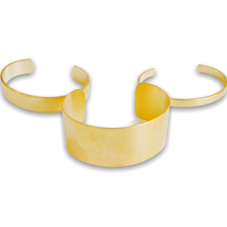 May include: Three gold-colored cuff bracelets. The bracelets vary in width, with one wide cuff and two thinner, open-ended bands. The jewelry is displayed on a white surface, showcasing the reflective quality of the metal.
