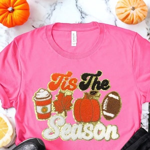 May include: A pink t-shirt with the text "Tis the Season" in gold glitter lettering. The shirt also features a pumpkin, a football, a coffee cup, and a leaf in orange and brown.