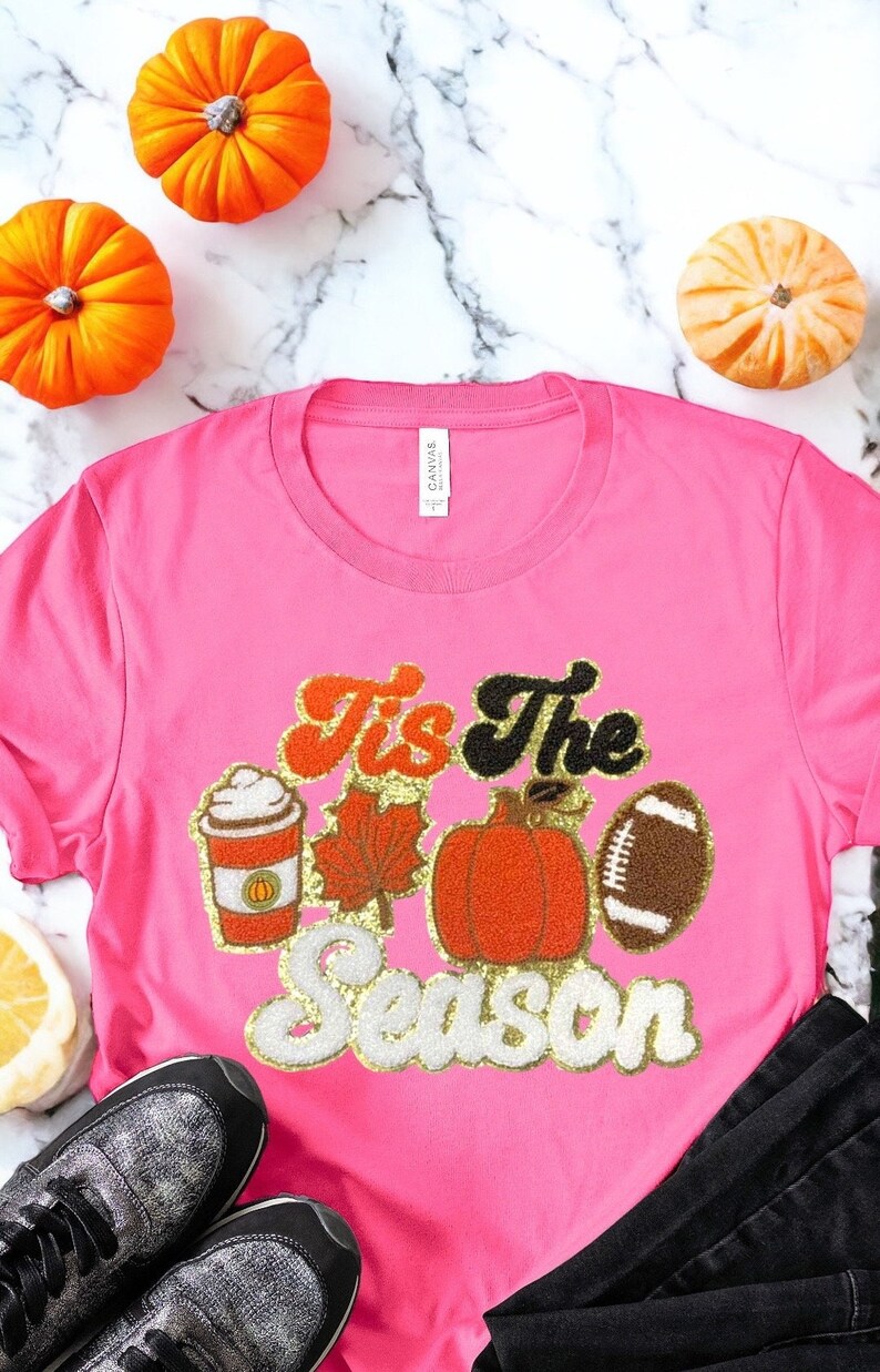 May include: A pink t-shirt with the text "Tis the Season" in gold glitter lettering. The shirt also features a pumpkin, a football, a coffee cup, and a leaf in orange and brown.