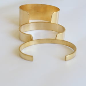 May include: Three gold-colored metal cuff bracelets of varying widths are arranged on a white surface. The bracelets have a simple, open design, with a gap at the wrist. The largest cuff is approximately 2 inches wide, the middle one is 1 inch wide, and the smallest is 0.5 inches wide.