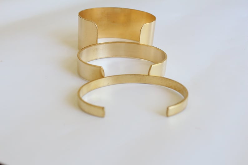 May include: Three gold-colored metal cuff bracelets of varying widths are arranged on a white surface. The bracelets have a simple, open design, with a gap at the wrist. The largest cuff is approximately 2 inches wide, the middle one is 1 inch wide, and the smallest is 0.5 inches wide.