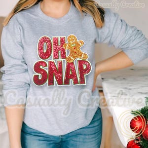 Puede incluir: Una sudadera gris con un diseño de lentejuelas rojas y doradas que dice "Oh Snap" con un hombre de jengibre en el medio.