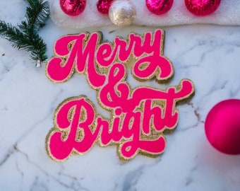 Peut inclure: Un panneau "Merry & Bright" rose et or avec une police rétro festive. Le panneau est en feutre et a une finition pailletée.