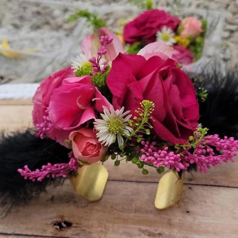 May include: A floral wrist cuff featuring vibrant pink roses, small white daisies, and delicate pink accents. The flowers are set on a gold-colored metal cuff with black feathers.