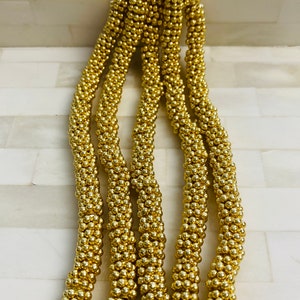 May include: A close-up of a strand of gold beads. The beads are small and round, and they are strung together in a tight, even pattern. The beads are shiny and reflect the light.