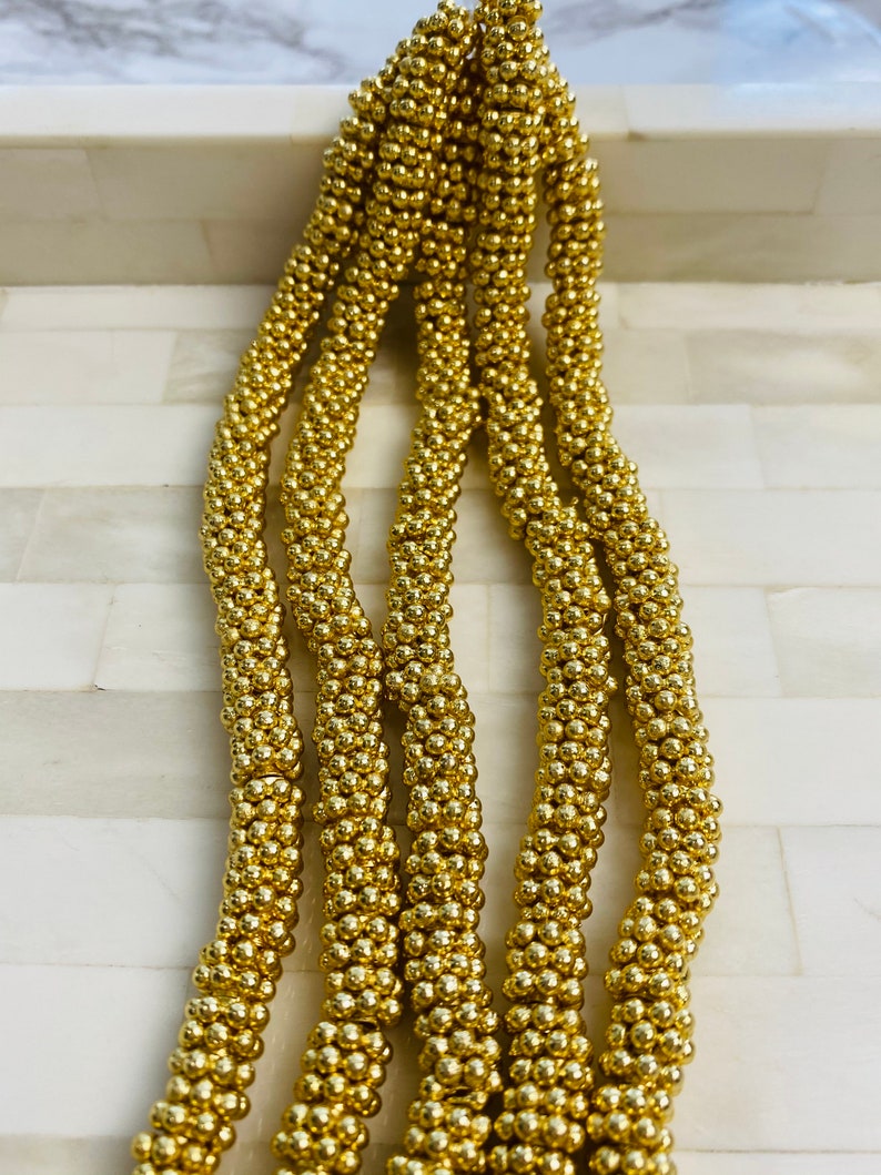 May include: A close-up of a strand of gold beads. The beads are small and round, and they are strung together in a tight, even pattern. The beads are shiny and reflect the light.