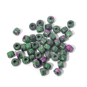 May include: A collection of green and magenta gemstone beads. The beads are cylindrical with a hole through the centre, and are scattered across a white surface. The gemstones have a mottled appearance, with a mix of green and magenta colours.