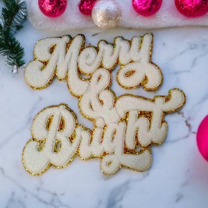 Op de afbeelding: Witte viltletters die "Merry & Bright" spellen met gouden glitteraccenten op een wit marmeren oppervlak.  Roze ornamenten zijn rondom de letters verspreid.