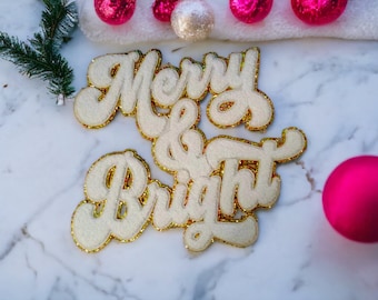 Peut inclure: Lettres en feutre blanc épellant "Merry & Bright" avec des accents de paillettes dorées sur une surface en marbre blanc. Des ornements roses sont éparpillés autour des lettres.