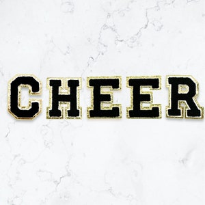 Op de afbeelding: Zwarte viltletters met gouden glitterrand die het woord "CHEER" spellen.