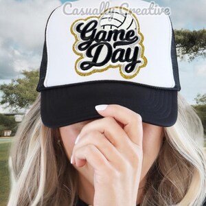 May include: A white and black trucker hat with a gold glitter patch that says "Game Day" and a white volleyball. The hat is worn by a person with blonde hair.